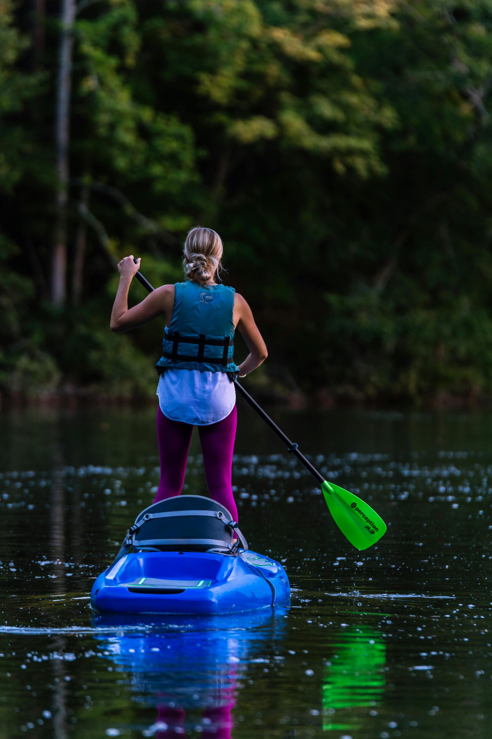 A woman enjoys stand-up kayaking on a calm lake surrounded by lush greenery, wearing a life vest.
